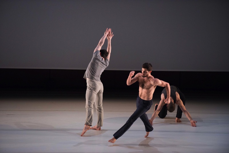 A trio of men dance center stage; each performing their own solo. One is kneeling on the ground, while the other is in mid-run and the other stretches both arms upwards toward the ceiling